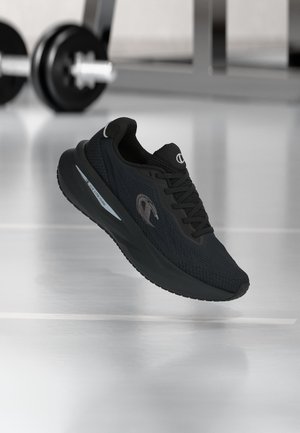 Schwarzer Sportschuh mit Schnürsenkeln und Markenlogo, der in Innenräumen auf einem reflektierenden Boden mit unscharfen Fitnessgewichten im Hintergrund schwebt.