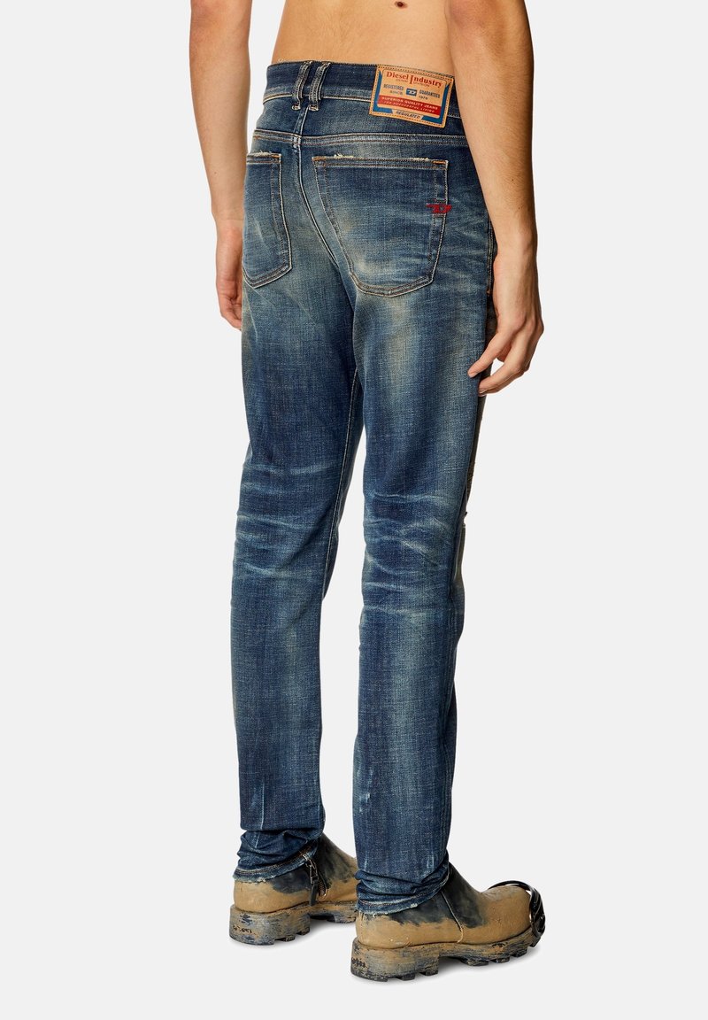 Blauwe denim jeans met een vervaagde, versleten uitstraling, vijf zakken en een leren merkpatch op de tailleband. Draagt met bruine werkschoenen.