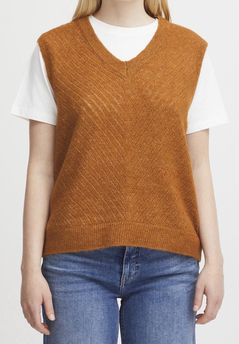 Braune Strickweste mit V-Ausschnitt, mit Chevron-Muster und geripptem Saum, getragen über einem weißen T-Shirt mit blauer Jeans.