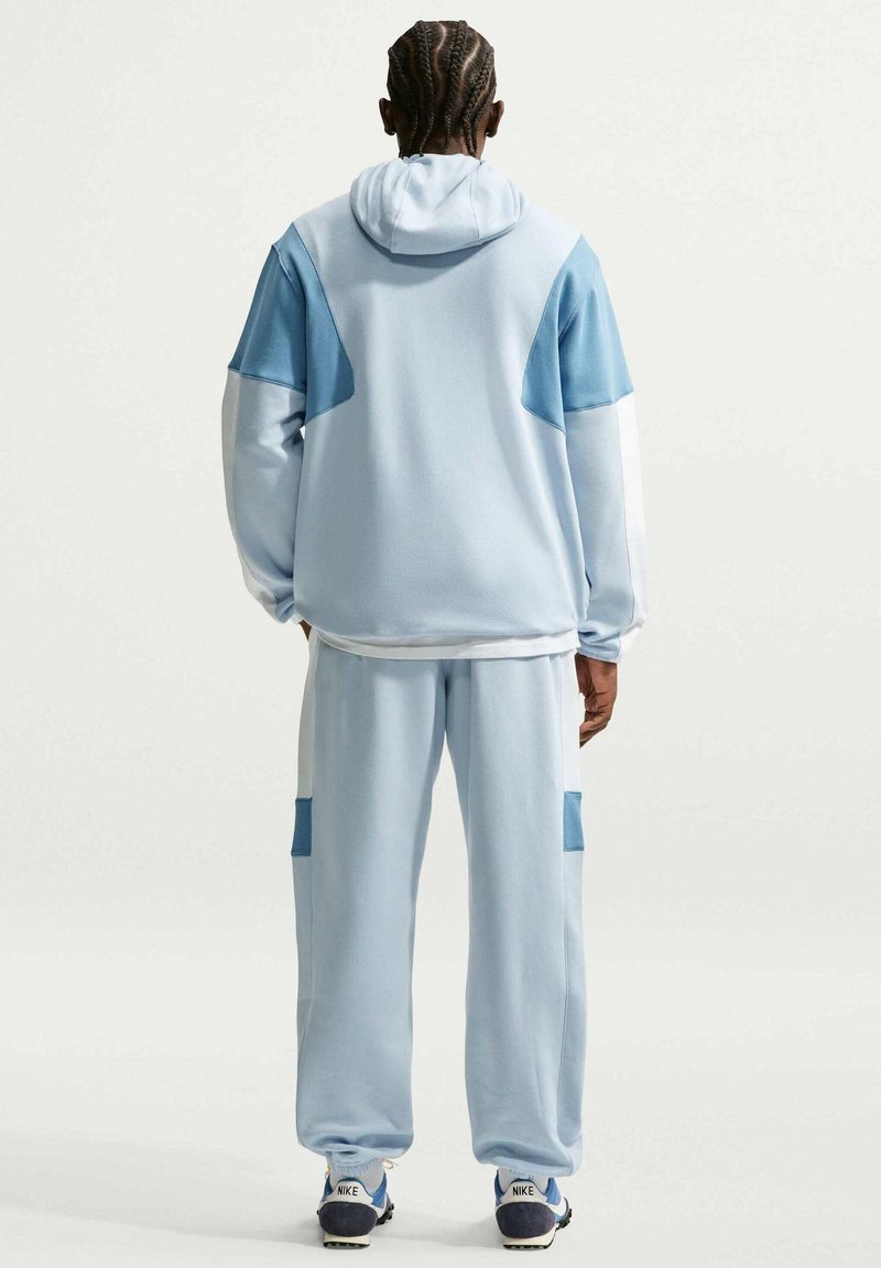 Persoon met gevlochten haar staat met de rug naar de camera, draagt een lichtblauw trainingspak met donkerblauwe vlakken en witte Nike-sneakers op een effen achtergrond.