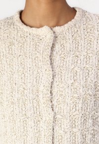 Cardigan di un leggero color crema texture con scollo rotondo e chiusura a un bottone, caratterizzato da un motivo intrecciato e materiale morbido e soffice.