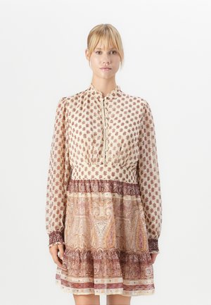 Kvinna klädd i en kräm- och brunmönstrad långärmad klänning med knappar och en blandad blommig och paisley-mönster, stående mot en vit bakgrund.