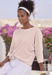 Maglione rosa chiaro con texture e maniche a tre quarti, abbinato a pantaloni bianchi e una cintura bianca. Accessoriato con orecchini a cerchio grandi.