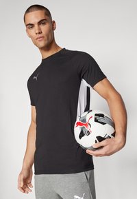 Tricou sportiv negru cu panouri laterale albe, având un mic logo gri. Pantaloni de trening gri. Ținând o minge de fotbal alb-negru cu accente roșii.