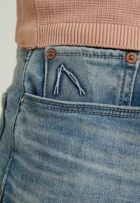 Ljust blå denimjeans med en synlig bakficka, med två metallknappar och en fransad detalj vid fickans kant. Texturerad tyg.