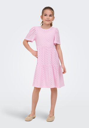 Jeune fille debout, main sur la hanche, portant une robe rose perforée et des chaussures beiges tissées à enfiler sur un fond uni.