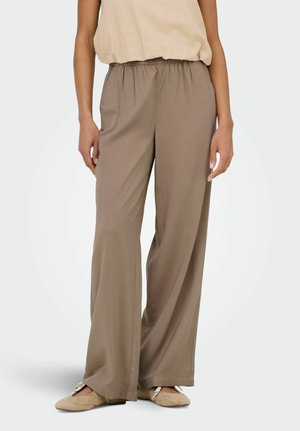 Femme portant un pantalon large taupe ample, un haut sans manches beige et des chaussures plates beiges, debout devant un fond uni.