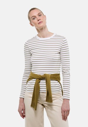 Femme portant une chemise blanche à manches longues avec de fines rayures marron et un pantalon beige, avec un pull vert olive noué autour de la taille.