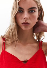 Rode mouwloze top met een V-hals, textuurstof en fijne spaghetti banden. Beschikt over een decoratieve kraal bij de halslijn voor extra detail.