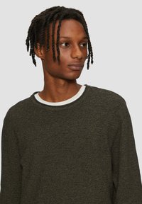 Dunkelgrüner Strickpullover mit strukturierter Oberfläche, Rundhalsausschnitt und langen Ärmeln, über einem weißen T-Shirt getragen.