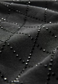 Černá tkanina s texturovaným vzorem tvořeným malými černými flitry uspořádanými v mřížkovém uspořádání, vytvářející jemný lesk.