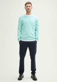 Mintgroene trui met V-hals, lichtgewicht textuur, gecombineerd met marineblauwe broek en zwarte veterschoenen. Model staat zelfverzekerd met handen in de zakken.