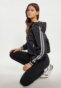 Sort vindjakke med hvide striber, lynlåslommer og justerbar hætte. Kombineret med sorte bukser og hvide sneakers.