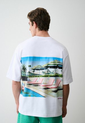 Mann trägt weißes T-Shirt mit bunter Poolszene mit Liegestühlen und Sonnenschirmen auf dem Rücken.