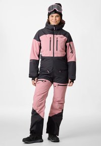 Tuta da sci con blocchi di colore nero e rosa chiaro, materiale impermeabile, due tasche anteriori con zip e cappuccio aderente, abbinata a occhiali da sci.