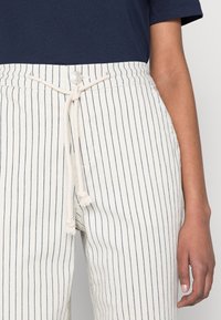 Pantalon à rayures blanches avec des lignes verticales bleu marine, doté d'une taille à cordon de serrage et d'une fermeture à bouton. Tissu en coton avec une texture lisse.