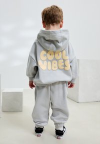 Completo con felpa con cappuccio e pantaloni della tuta grigi, con la scritta "COOL VIBES" in giallo sulla schiena, polsini a coste e scarpe da ginnastica nere con dettagli bianchi.
