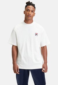 Biała bawełniana koszulka T-shirt z krótkim rękawem i okrągłym dekoltem, z małym niebieskim, czerwonym i czarnym logo FILA po lewej stronie klatki piersiowej. Prosty design.