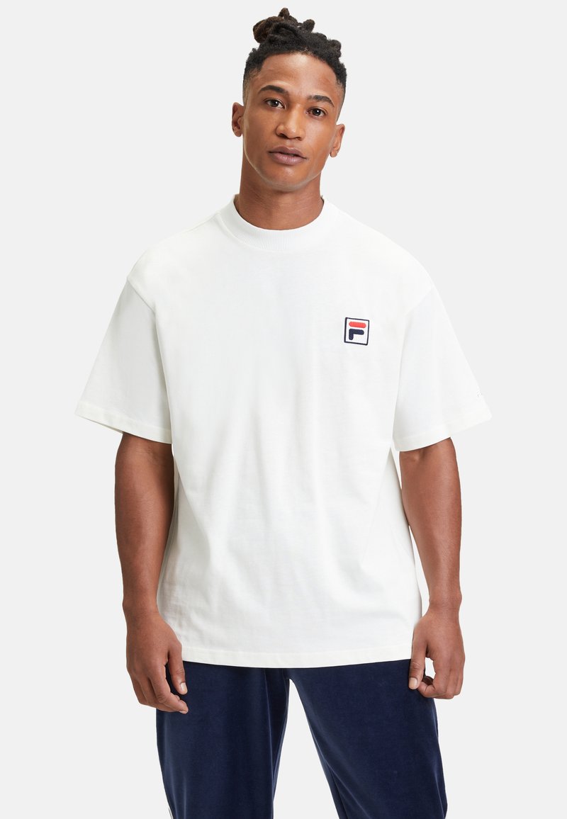 Biała bawełniana koszulka T-shirt z krótkim rękawem i okrągłym dekoltem, z małym niebieskim, czerwonym i czarnym logo FILA po lewej stronie klatki piersiowej. Prosty design.