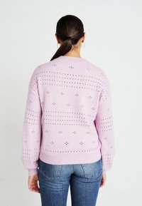 Pulover tricotat de culoare roz deschis, cu un model de mici găuri, manșete rigate și decolteu tip echipaj, purtat cu blugi din denim albastru.