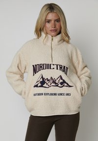 Kvinna i beige sherpa fleece med berggrafik och texten "NORDIC TRAIL" och "OUTDOOR EXPLORING SINCE 1953," med händerna i fickorna.