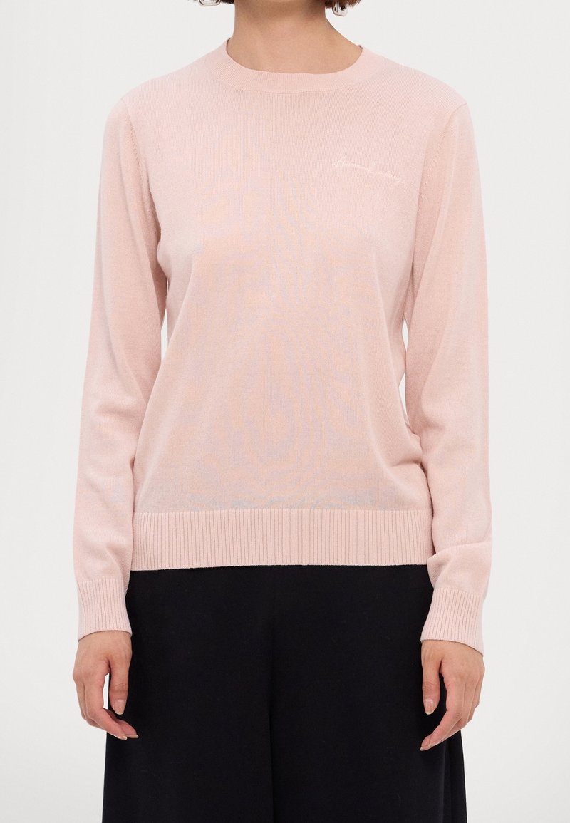 Femme portant un pull en tricot à manches longues rose clair avec des poignets et un ourlet côtelés, associé à un pantalon noir ample.