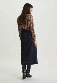 Jupe longue en denim bleu foncé avec des coutures jaunes, associée à un blouse à motifs noirs et rouges. Texture lisse, silhouette ajustée.