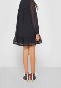 Námořnická modrá průsvitná šaty s volánkovým lemem a dlouhými rukávy, s jemným vzorem. Doplněno vícebarevnými vysokými teniskami.