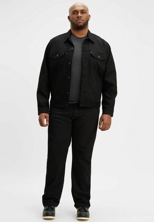 Chaqueta de mezcla de algodón negra con dos bolsillos frontales y cierre de botón, combinada con una camiseta negra y pantalones negros a juego, completada con calzado negro.