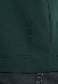 Mörkgrön kortärmad skjorta med texturerad tyg, som har tre små fyrkantiga detaljer i rad på den nedre sidan.