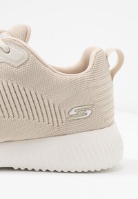 Beige idrottssko med en texturerad mesh-överdel, en tjock vit sula och subtila ribbade detaljer på hälen med en logotyp.