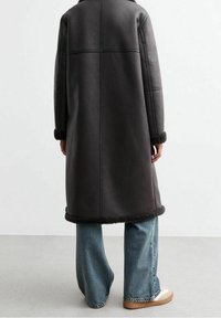 Cappotto lungo in pelle nera con rifiniture in shearling ai polsini e all'orlo, mostrato di spalle con jeans a gamba larga e calzature beige.