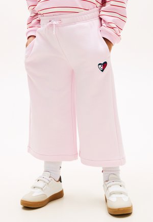 Barn iført lyserøde vidde bukser med snøre i taljen og hjertepatch, kombineret med hvide sneakers og stribet langærmet top.