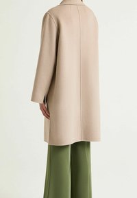 Cappotto beige in misto lana con maniche lunghe, taglio dritto, cucitura centrale sulla schiena e spacchi laterali, abbinato a pantaloni verdi a gamba larga.