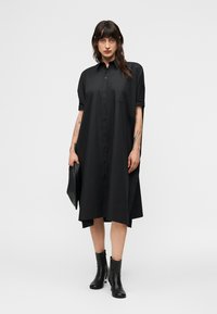 Γυναίκα που φοράει ένα φαρδύ μαύρο φόρεμα με κουμπιά, μαύρα μποτάκια αστραγάλου, κρατώντας ένα μαύρο τσαντάκι χειρός, στέκεται μπροστά σε λευκό απλό φόντο.