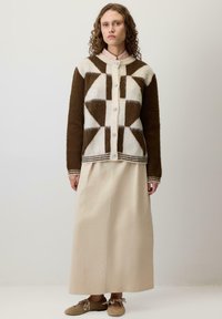 Kobieta w brązowym i białym kardiganie w geometryczny wzór, beżowej długiej spódnicy oraz beżowych płaskich butach z kokardkami, stojąca na jednolitym tle.