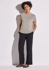 Žena s kudrnatými vlasy stojí proti jednoduché zdi, oblékla si světle šedý top s krátkým rukávem, černé rovné kalhoty a černé ploché boty, ruce má v kapsách.