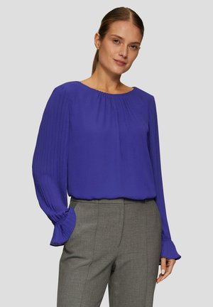 Lila Bluse mit langen Ärmeln und plissierten Akzenten, runder Ausschnitt, kombiniert mit grauen, taillierten Hosen. Glatter Stoff, entspannte Passform.