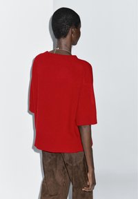 Czerwona wełniana sweter z krótkimi rękawami i prążkowanym wykończeniem, w połączeniu z brązowymi zamszowymi spodniami, widziany z tyłu na neutralnym tle.