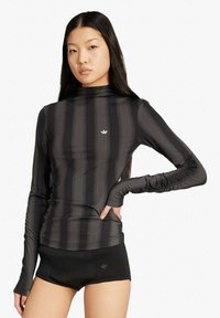 Donna con capelli lunghi e neri, indossa una maglia a maniche lunghe a righe nere e grigio scuro e pantaloncini sportivi neri con loghi.