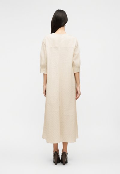 Γυναίκα με μακριά σκούρα μαλλιά, φορώντας μπεζ φόρεμα μέτριου μήκους με μανίκια τριών τετάρτων και καφέ παπούτσια με τακούνι, στέκεται με την πλάτη στραμμένη.