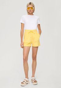 Geel katoenen shorts met een trekkoord in de taille en twee voorzakken, gecombineerd met een witte t-shirt en stevige witte sandalen. Grote gele zonnebril.