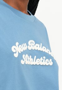 T-shirt de algodão azul claro com letras 3D brancas que dizem "New Balance Athletics", com decote redondo e mangas curtas.