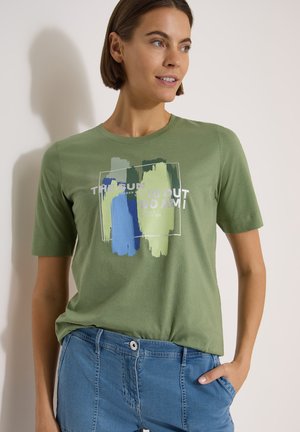 Kvinde iført olivengrøn t-shirt med blå og grøn penselstrøg-design og afslappede blå jeans, stående med hånden i lommen.