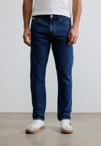 Μπλε τζιν παντελόνια με ίσια γραμμή, πέντε τσέπες, θηλιές για ζώνη και κλείσιμο με κουμπί, σε συνδυασμό με λευκά αθλητικά παπούτσια. Ομαλή υφή.