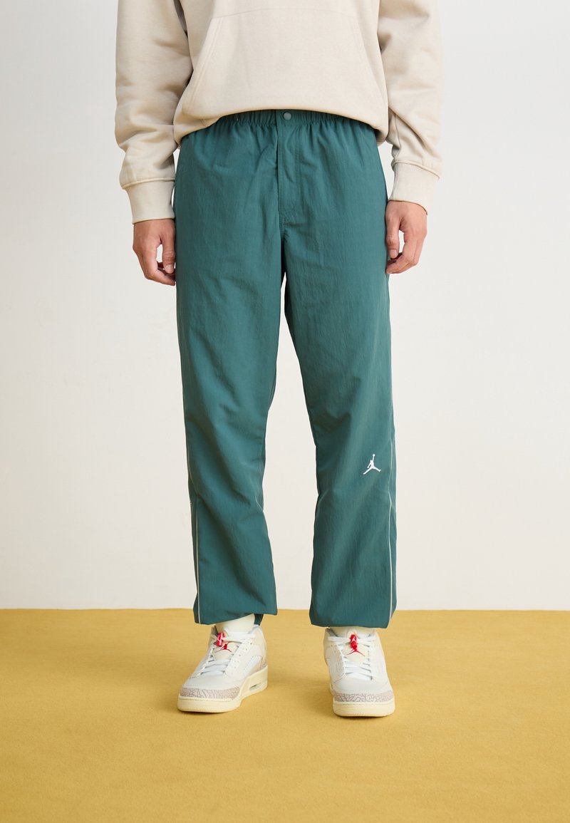 Zelené lehké kalhoty s elastickým pasem a bočními pruhy, s bílým logem Jordan na levé noze, kombinované s bílými teniskami.