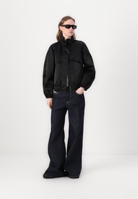 Black cropped bomber jacket with elastic waistband and cuffs; paired with wide-legged dark denim jeans; model wears black sunglasses.