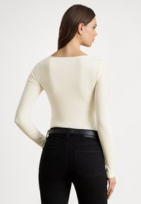 Top a maniche lunghe a costine in crema chiaro con scollo quadrato, abbinato a jeans neri a vita alta con cintura nera.