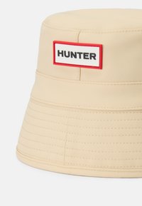 Klobouk z lehkého krémového materiálu. Obsahuje červenou a bílou nášivku s nápisem "HUNTER" v tučném černém písmu. Prošité detaily.