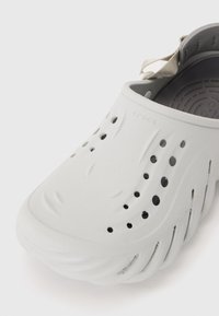Białe klapki Crocs z teksturowaną powierzchnią, otworami wentylacyjnymi i paskiem na piętę. Charakteryzują się gładkim, elastycznym materiałem.